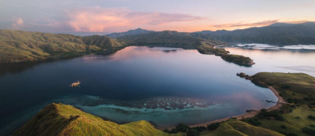 beautiful image from indonesia. A serene landscape featuring a calm lake surrounded by green hills, with a boat near the shore against a colorful sunset sky.