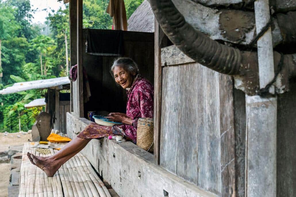 An elderly woman sits outside a rustic wooden home, smiling while holding a bowl, with lush greenery in the background.