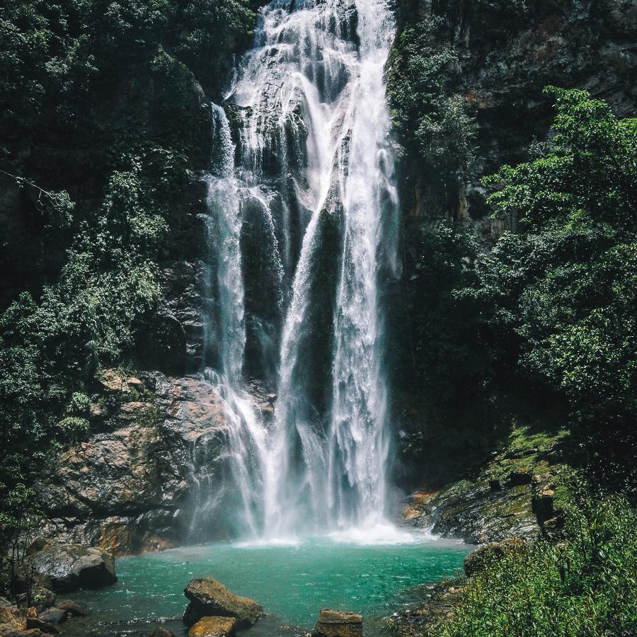 CuncaRami. waterfall. Lush green landscape surrounding a tall, cascading waterfall into a turquoise pool below. Sunlight filters through trees.