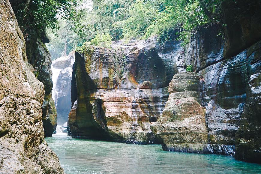 CuncaWulang. waterfall and rocks. A serene canyon featuring tall, rocky cliffs and a flowing turquoise river beneath a lush green canopy.