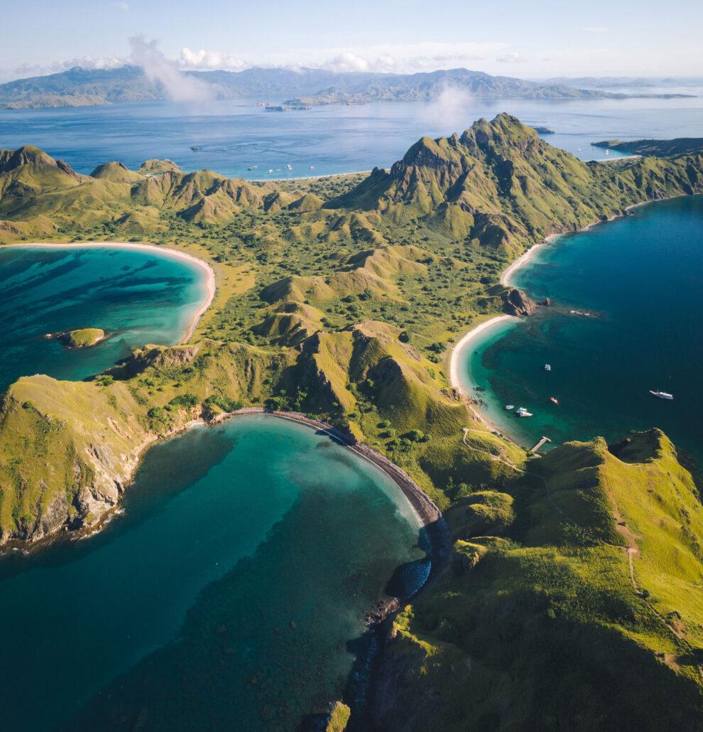 3 Days Komodo Dragon Island Boat Tours. Aerial view of lush green hills merging into turquoise waters and sandy beaches, with distant mountains under clear blue skies.