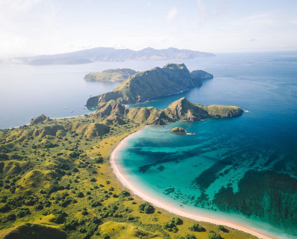 Aerial view of a lush green island with sandy beaches and clear turquoise waters, surrounded by distant mountains.