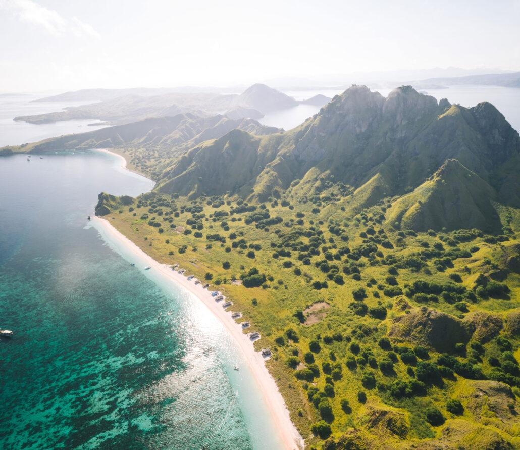 1 day komodo boat tour. Aerial view of lush green hills meeting a sandy beach and turquoise waters, with distant islands on the horizon.
