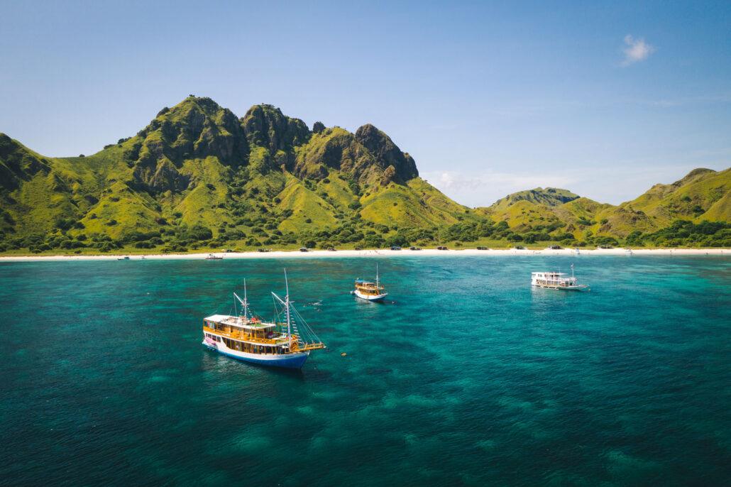 Three boats anchored in clear turquoise waters near a lush green coastal landscape and rugged mountains under a blue sky.