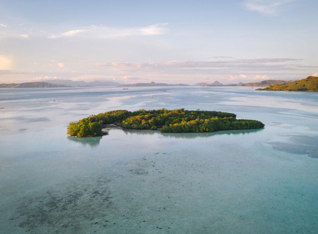 Aerial view of a lush green island surrounded by turquoise waters and distant mountains under a clear sky.