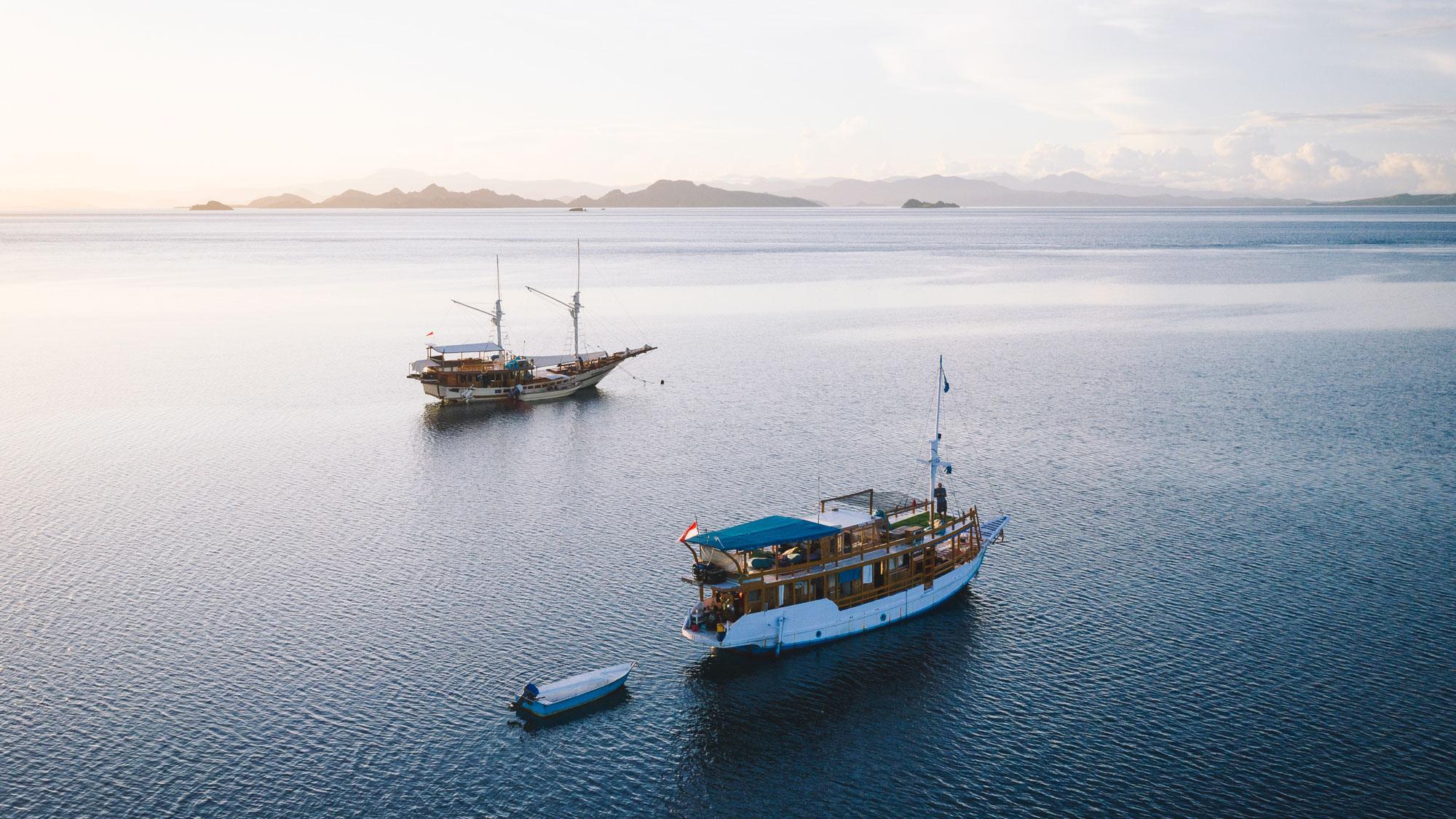 Komodo Boat Tour. Two wooden boats float on calm waters at sunset, with distant islands and a golden sky in the background.