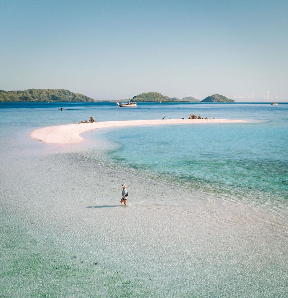 Komodo Boat Tour. A serene beach with a pink sandbar, turquoise waters, and distant green islands; a lone person walking through shallow water.