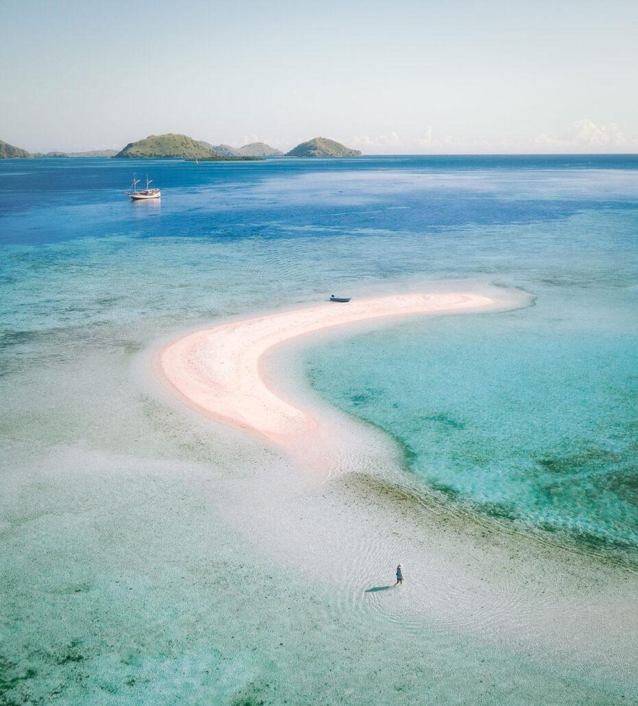 Komodo Boat Tour. Aerial view of a serene beach with a curved sandbar, clear turquoise waters, and a distant boat on the horizon.