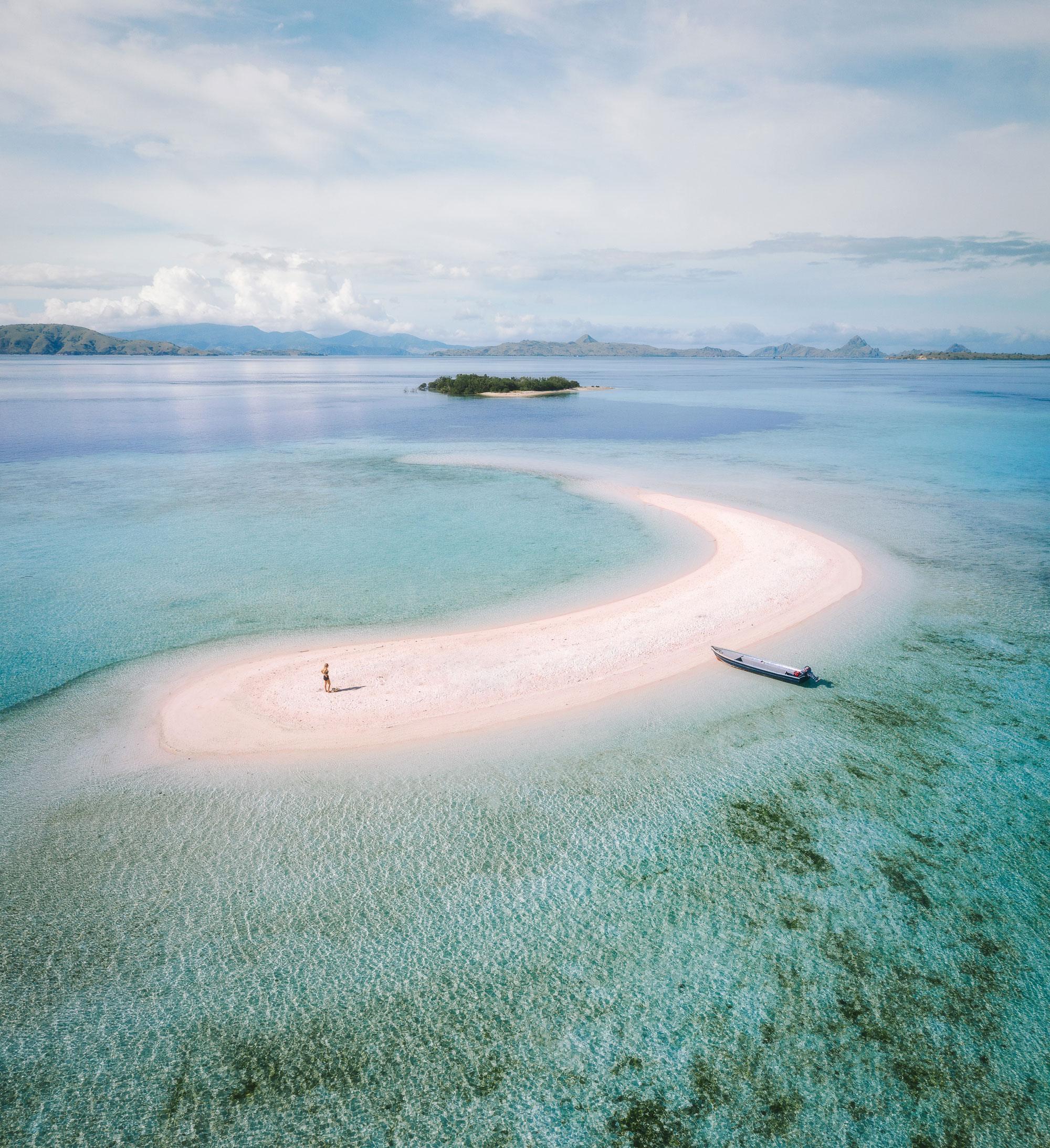 Komodo Boat Tour. Aerial view of a curved sandy beach on a turquoise lagoon, with a small boat and distant islands under a clear sky.