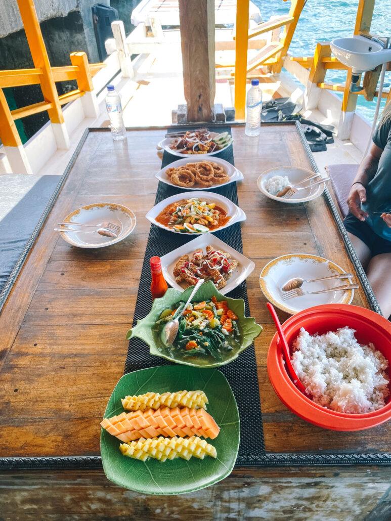 A wooden table on a boat displays various dishes, including seafood, rice, and tropical fruit, with ocean views in the background.