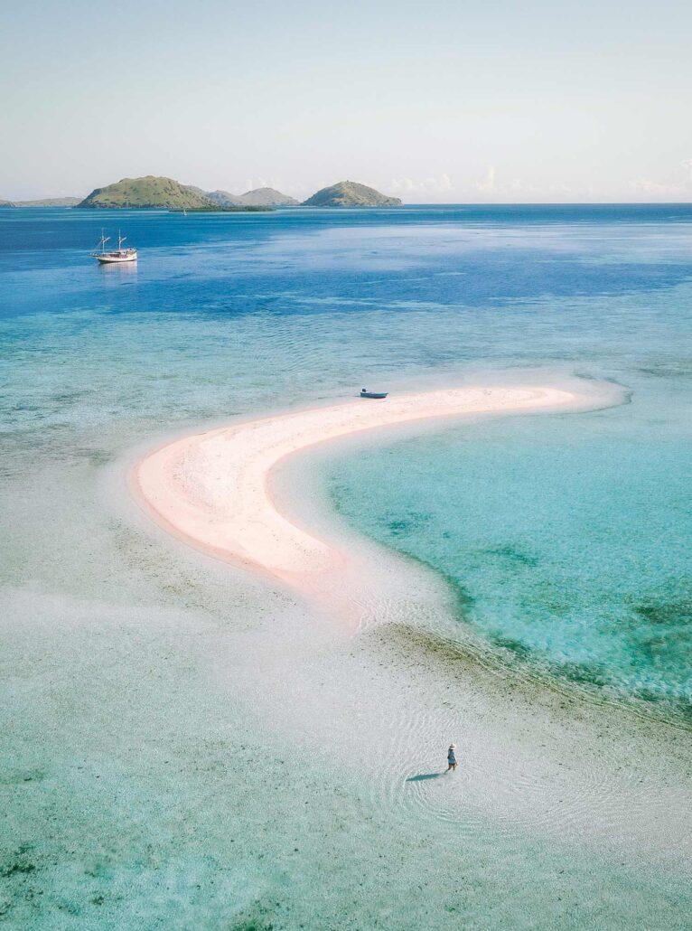 Takak Makassar. Aerial view of a sandy beach on a turquoise lagoon with a lone figure walking in shallow water and a ship in the distance.