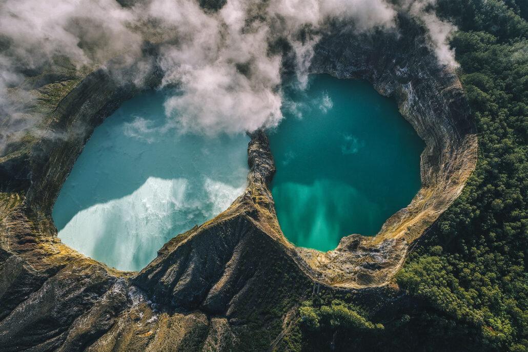 KelimutuItinerary - 2 vulcanoes. Aerial view of two vibrant turquoise lakes surrounded by lush green terrain and misty clouds.