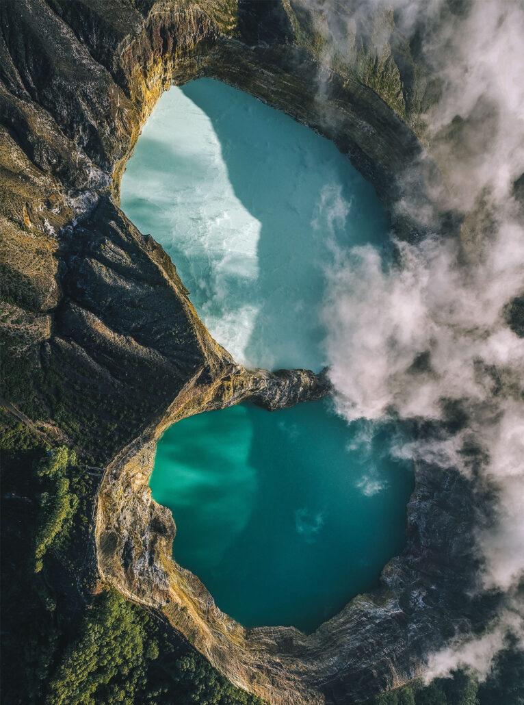 KelimutuItinerary - 2 vulcanoes. Aerial view of two volcanic lakes with turquoise water, surrounded by rugged cliffs and wispy clouds.