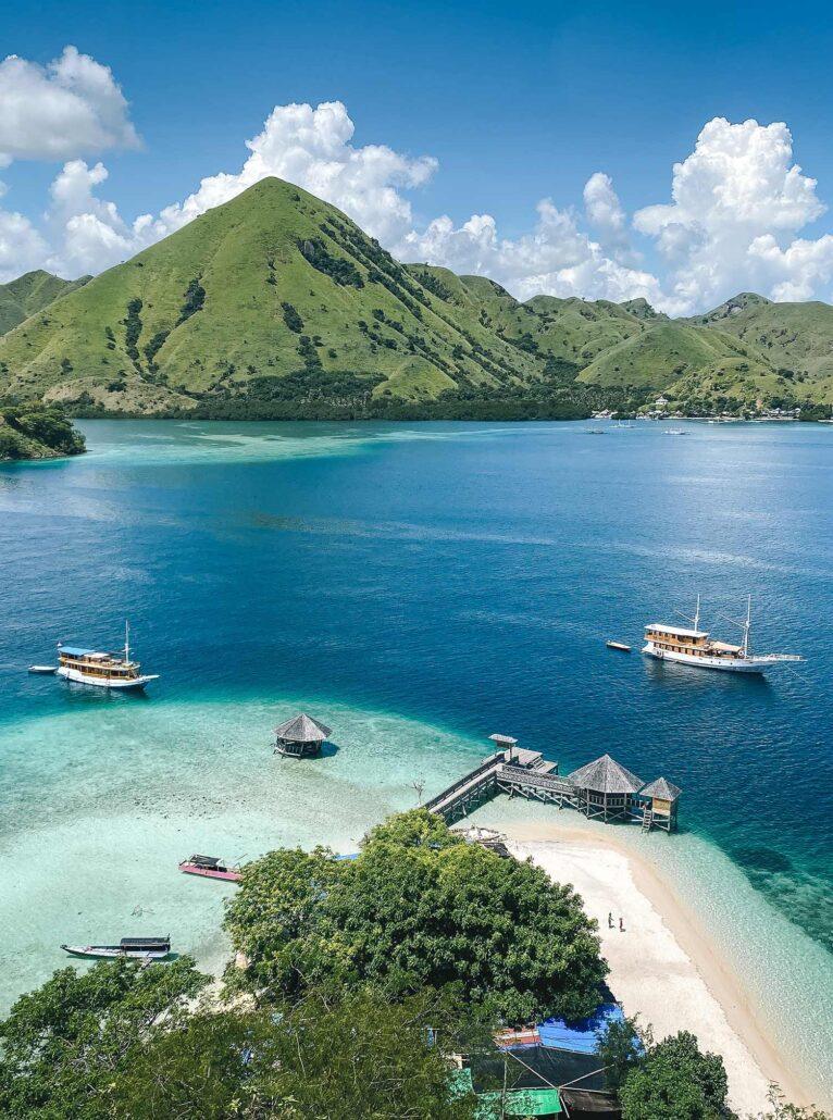 sandy beach on komodo island. A serene coastal scene with boats docked by a sandy beach, surrounded by lush green mountains and a clear blue sky.