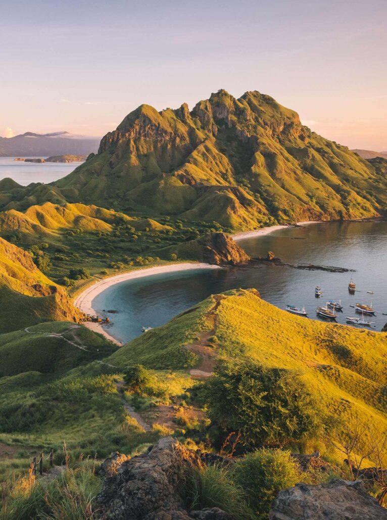 Padar island. Lush green hills surround a serene bay with a sandy beach and anchored boats, illuminated by the soft glow of sunset.