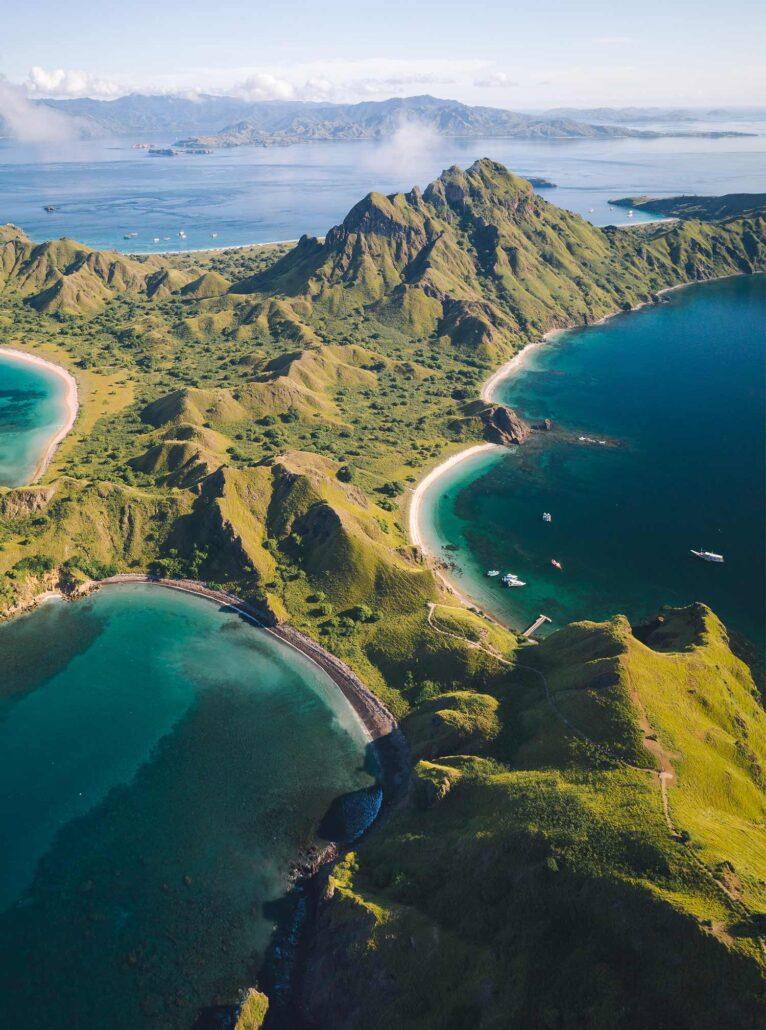 Padar island. Aerial view of green hills and two turquoise bays with white sandy beaches, surrounded by calm blue waters and distant mountains.