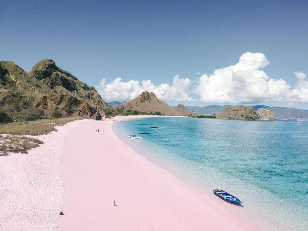 Pink beach. Aerial view of a pink sand beach, turquoise water, distant hills, and a few boats under a clear blue sky with white clouds.