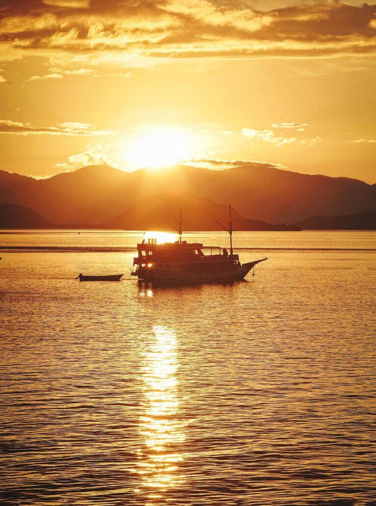 2 Days Komodo Boat Tour. Sunset in Indonesia. A silhouette of a boat on calm water at sunset, with mountains in the background and vibrant golden light reflecting on the surface.
