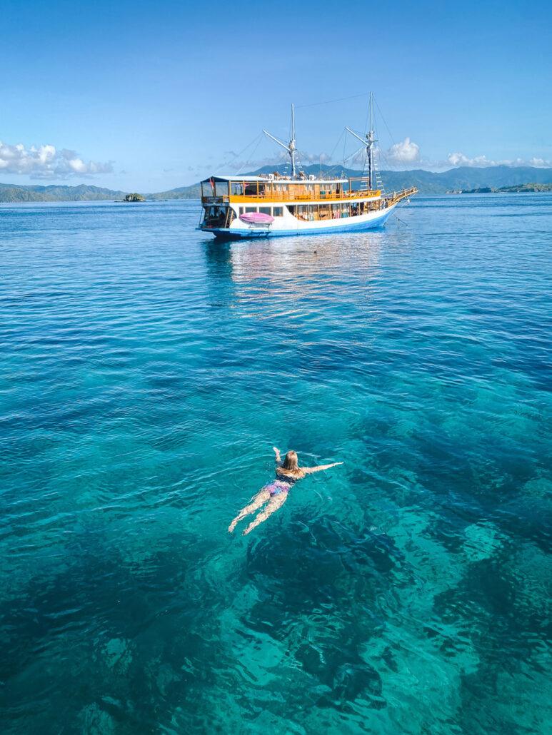 A person swims in clear turquoise water near a large wooden boat under a bright blue sky.