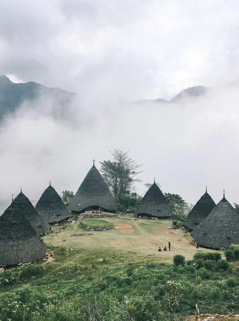 WaeRebo village. A serene landscape featuring traditional thatched-roof huts nestled in lush green hills, surrounded by misty mountains.