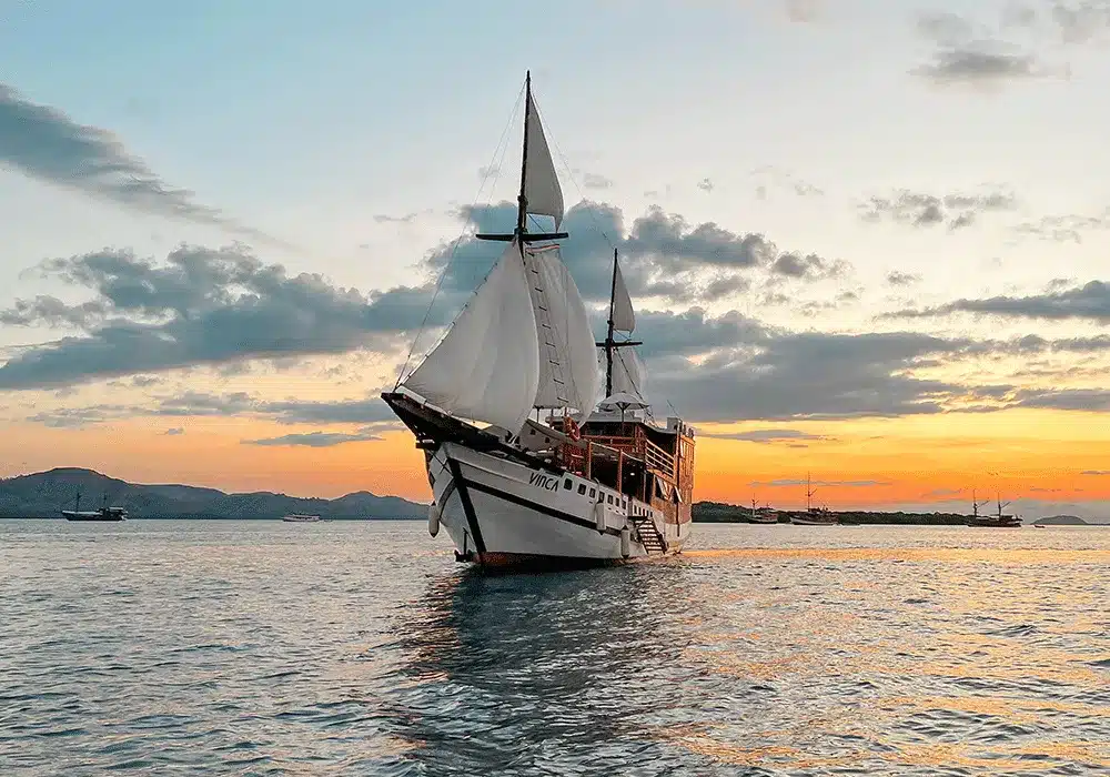 3 days shared trip. A majestic sailing ship with white sails glides through calm waters at sunset, surrounded by distant hills and dramatic clouds.