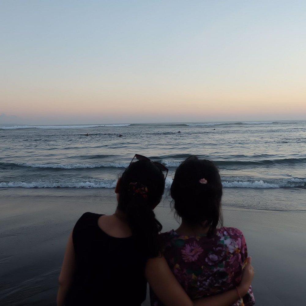 Batu Bolong. Two women standing together on a beach at sunset, gazing at the ocean waves and surfers in the distance.