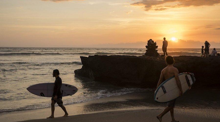 Batu Bolong. Two surfers walking along the beach at sunset, with silhouettes of people on rocks and waves in the background.