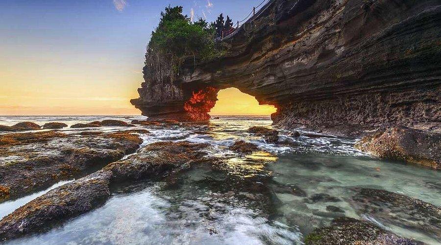 Batu Bolong. Scenic coastal scene featuring a rock arch at sunset, surrounded by water, rocky shores, and lush greenery.