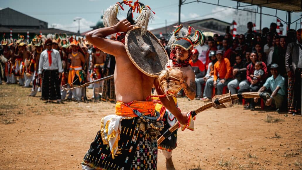 Caci traditional dance. Two dancers perform in traditional attire, wielding weapons, while an audience watches in a vibrant outdoor setting.