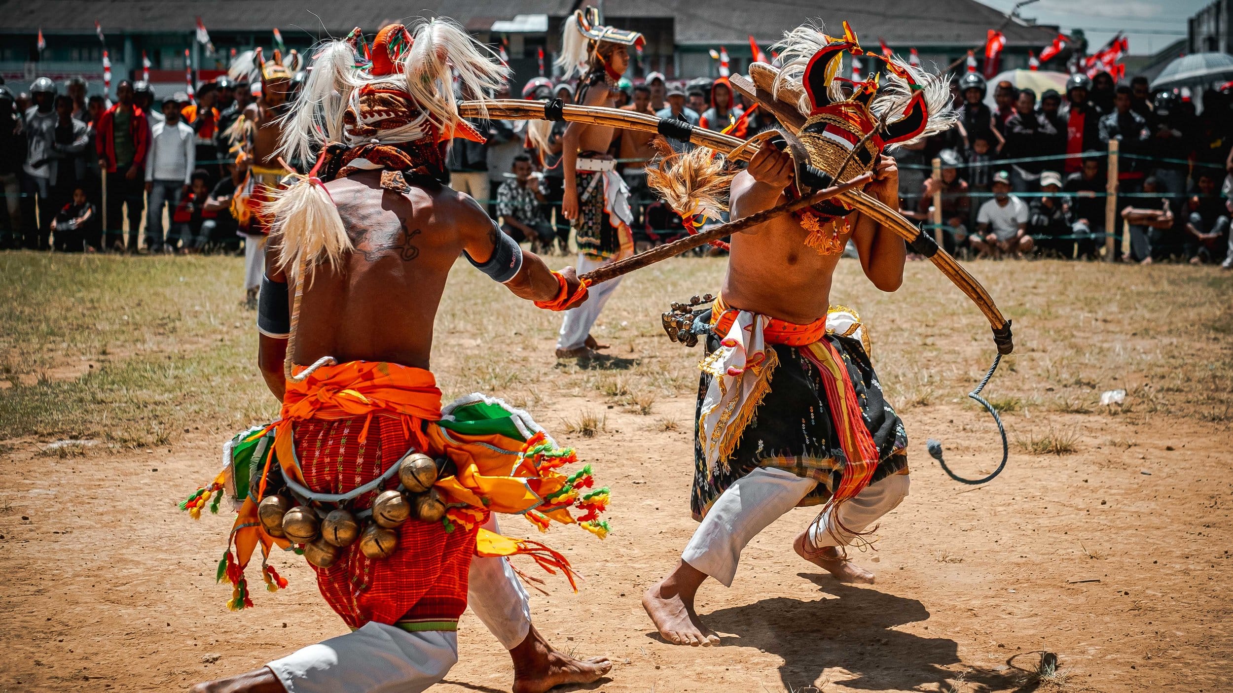 Caci traditional dance. Two dancers in traditional attire perform in a vibrant display, wielding bows against a backdrop of an enthusiastic crowd.