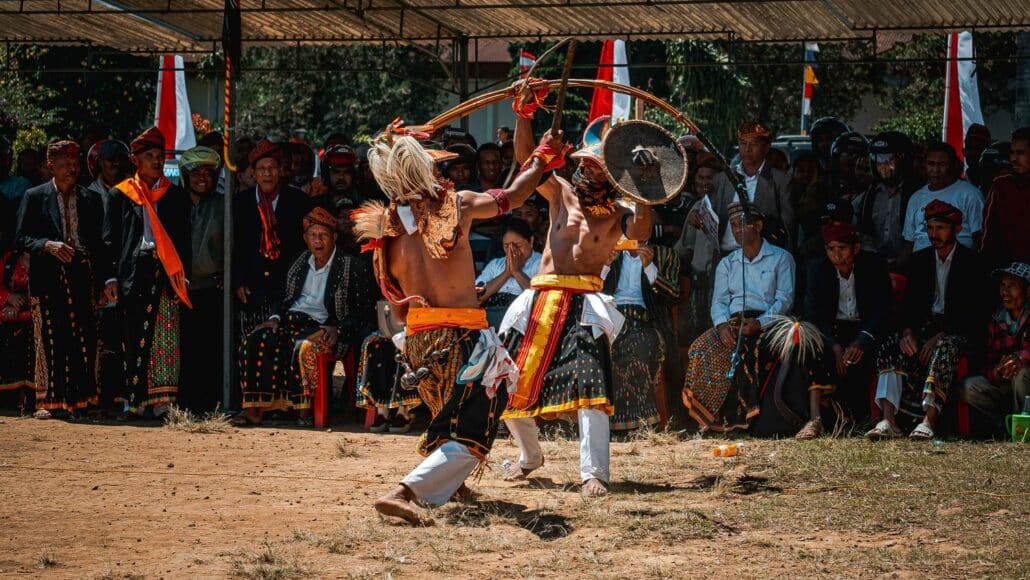 Caci traditional dance. Two dancers in traditional attire engage in a dynamic dance battle, surrounded by an audience in colorful clothing.