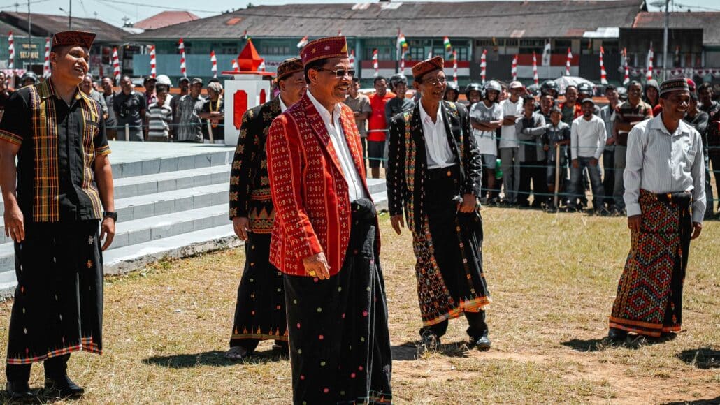 Caci traditional dance. Men in traditional attire stand in a grassy area, with spectators and colorful decorations in the background.
