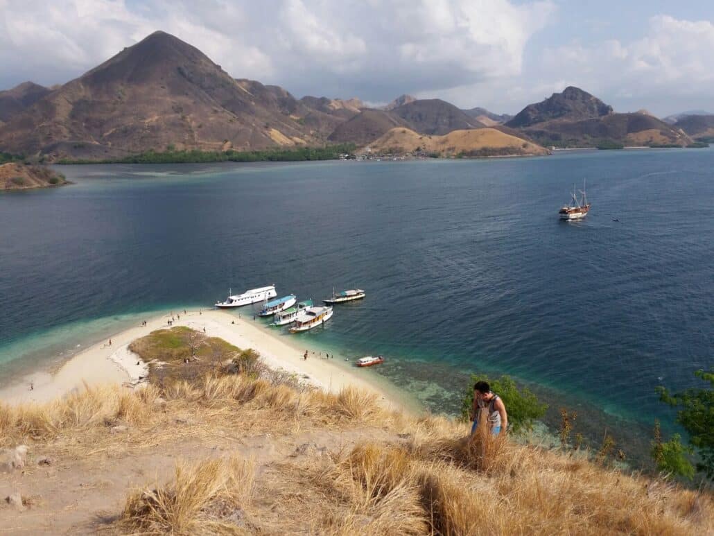 Flores Komodo boat tour. A view of a tropical bay with boats near a sandy beach, surrounded by lush green hills and distant mountains.