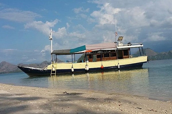 Flores Komodo boat tour. A colorful boat anchored on calm waters, with sandy shores and mountains in the background under a cloudy sky.