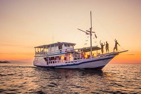 Flores Komodo boat tour. A traditional boat sails on calm waters at sunset, with two people waving from the deck under a vibrant sky.