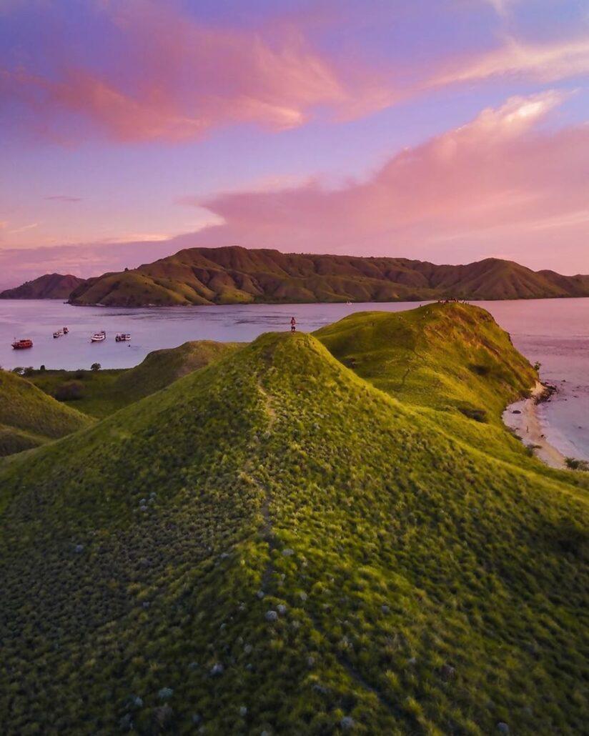 Gili Lawa Hiking. Sunset view over lush green hills and tranquil waters with anchored boats and vibrant purple-pink sky in the background.