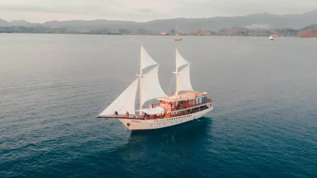 Komodo luxury tour. A large white sailboat with two sails navigates calm blue waters, surrounded by distant mountains and a hazy sky.