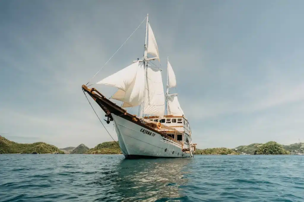 Komodo luxury tour. A white sailing ship with tall sails floats on calm blue waters, surrounded by green hills under a clear sky.