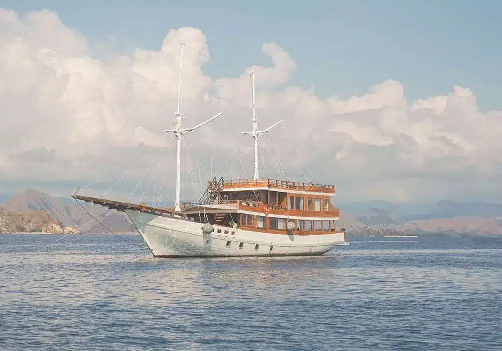 Komodo luxury tour. A wooden sailboat with two masts glides through calm waters, set against a backdrop of mountains and fluffy clouds.