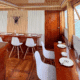 3 days shared trip. Cozy dining area on a boat, featuring wooden furniture, white chairs, and a scenic view of water through large windows.