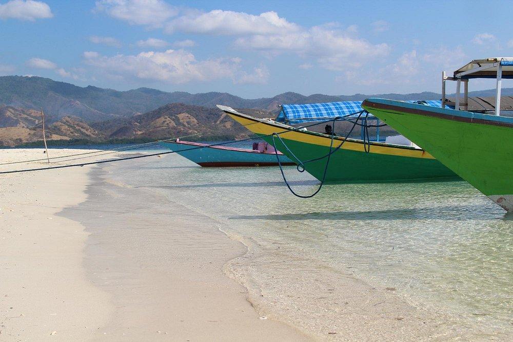 Riung 17 Islands Marine Park. Colorful boats anchored on a sandy beach with clear water, mountains in the background, and a blue sky with clouds.