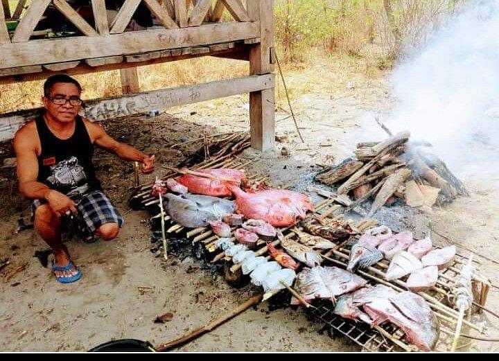 Riung 17 Islands Marine Park. A man sits beside a large grill, roasting various types of fish over an open flame in a rustic outdoor setting.