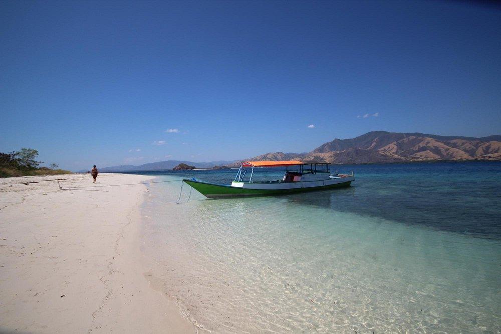 Riung 17 Islands Marine Park. A colorful boat anchored near a sandy beach, clear blue water, and distant mountains under a bright sky.