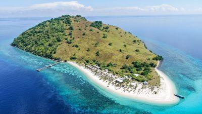 Sabolo Island, Labuan Bajo. Aerial view of a lush green island surrounded by turquoise waters and a sandy beach, with boats near the shore.
