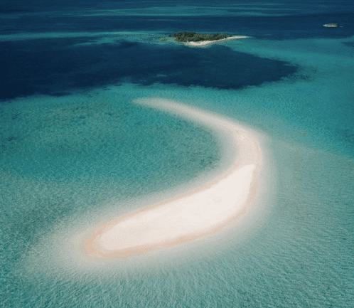 Takak Makassar - Aerial view of a curved sandy beach surrounded by clear turquoise water, leading to a small island in the distance.