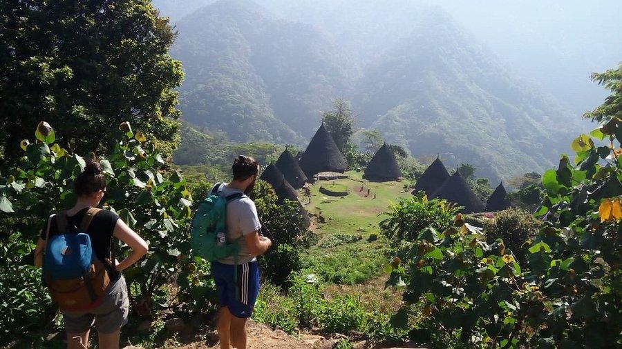Wae Rebo Village. Two hikers overlook a valley with cone-shaped huts surrounded by lush green mountains and trees.