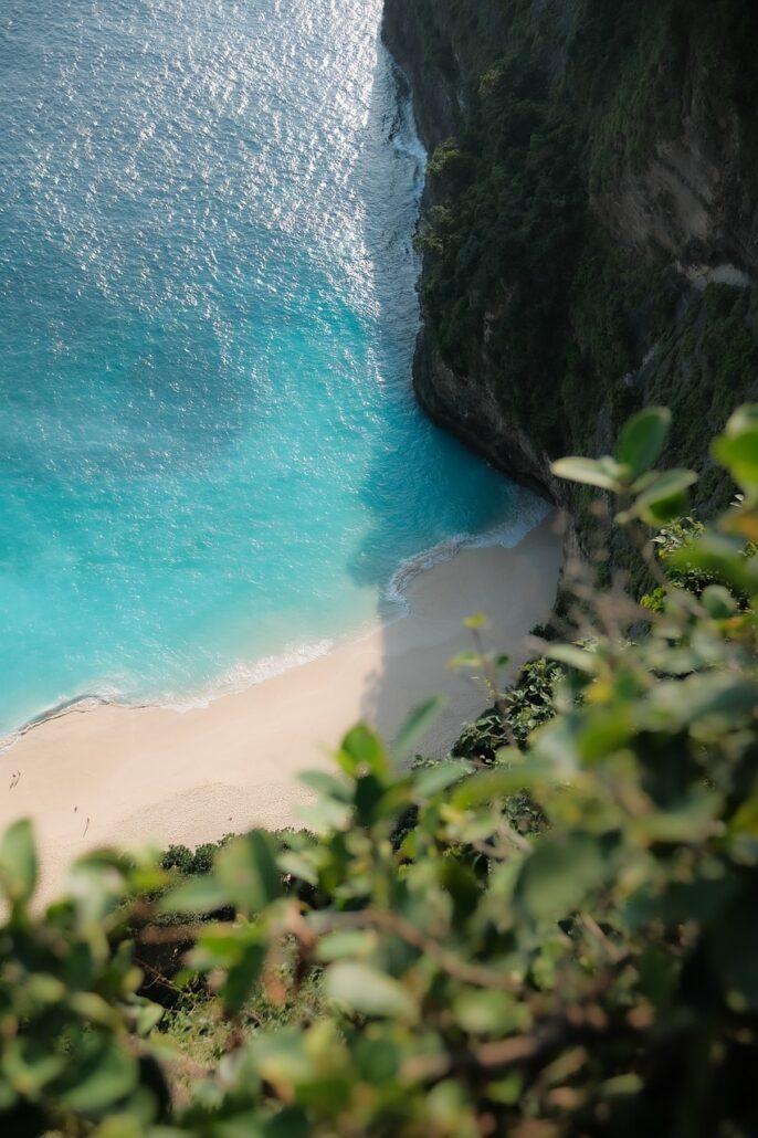 Bali Boat Tour. Aerial view of a serene beach with turquoise waters, sandy shore, and lush green cliffs framing the coastline.