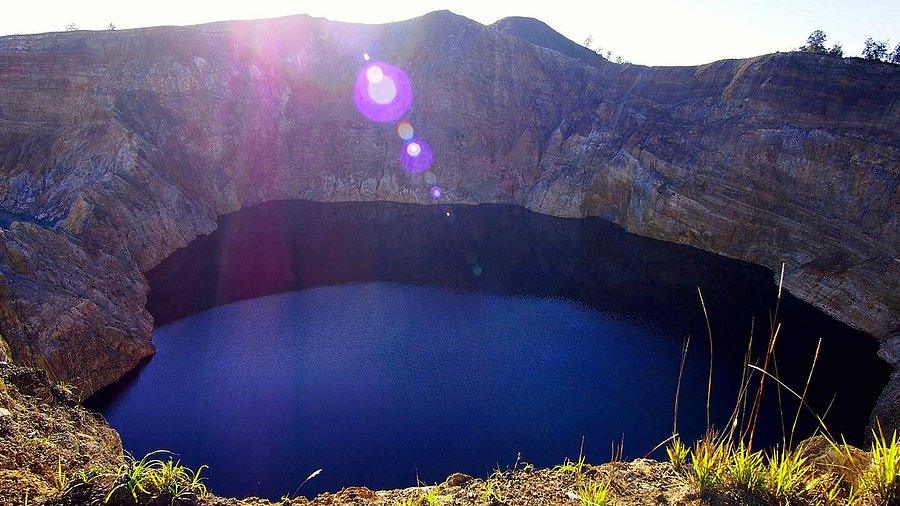 Kelimutu Volcano indonesia​, three colord volcano. A deep blue crater lake surrounded by rocky cliffs under bright sunlight with lens flare effects.