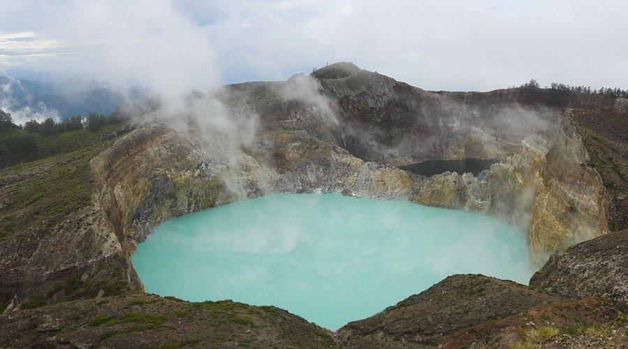 Kelimutu Volcano indonesia​, three colord volcano. A striking turquoise volcanic crater surrounded by rocky terrain and steam drifting from the edges under a cloudy sky.