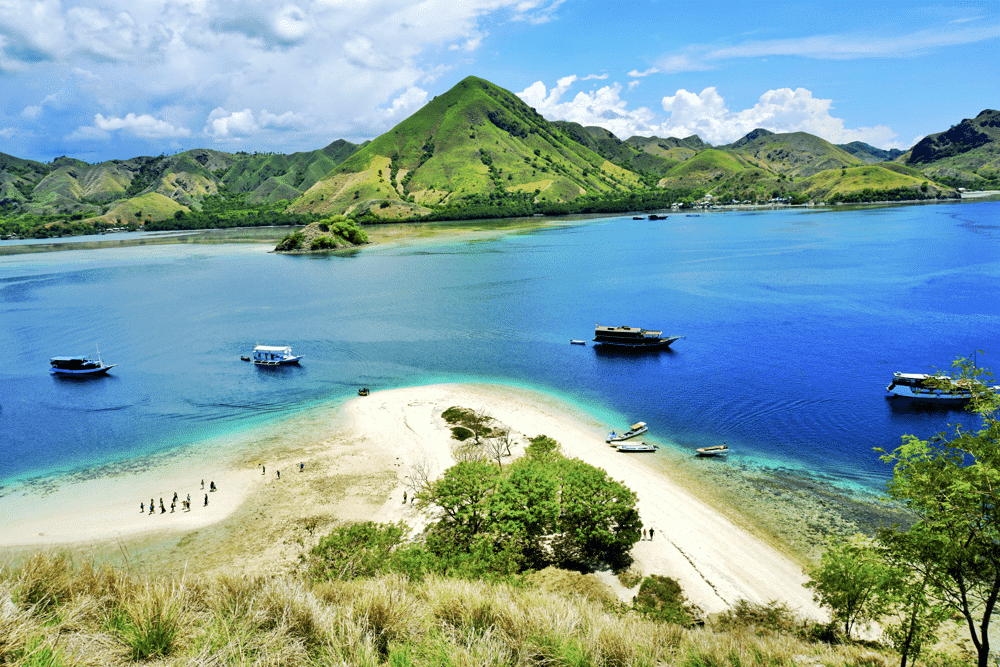 Kelor Island. Scenic tropical island view with sparkling blue waters, boats, sandy beach, and lush green hills under a bright sky.